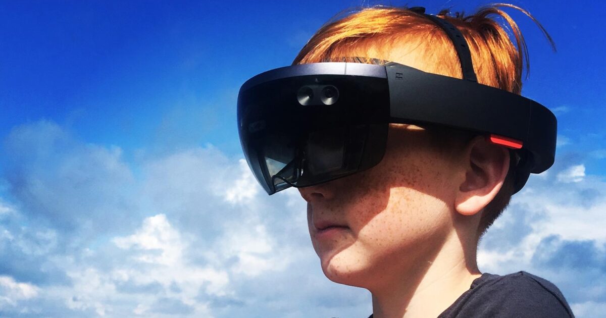 En ung person med rött hår bär ett stort, svart augmented reality-headset utomhus under en klarblå himmel med spridda moln.