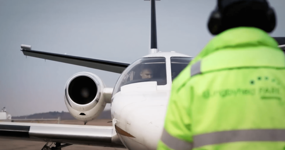 Ett litet vitt jetplan står parkerat på en landningsbana, med en person i neongulgrön varseljacka och svarta hörselkåpor i förgrunden.