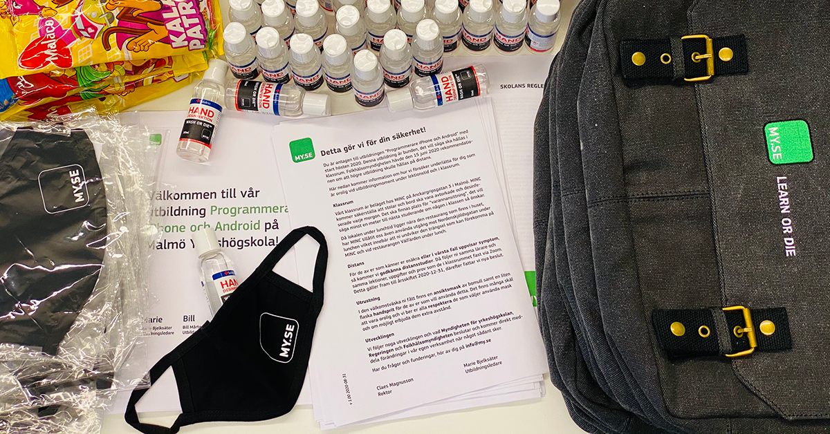 Ett bord med en svart ryggsäck, en ansiktsmask märkt MSE, flaskor med handsprit, papper på svenska, ett välkomstbrev och godispåsar, arrangerade för ett evenemang eller välkomstpaket.