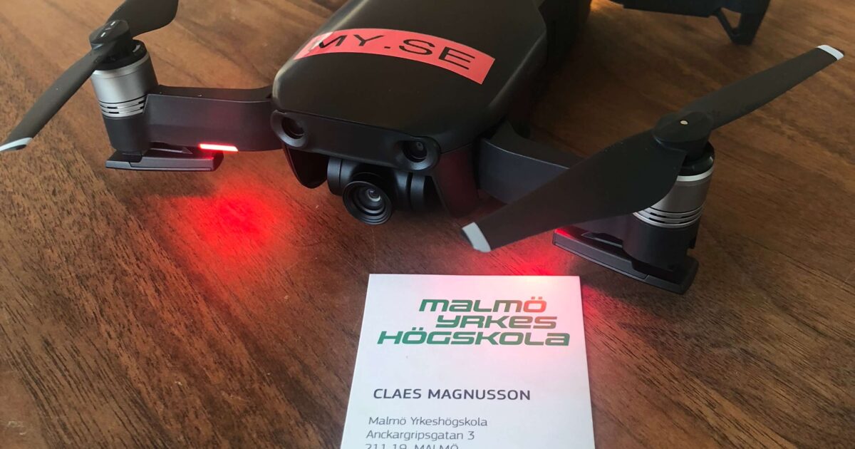 En liten svart drönare med röda lampor står på ett träbord bredvid ett visitkort för Malmö Yrkeshögskola och visar kontaktuppgifter till Claes Magnusson.