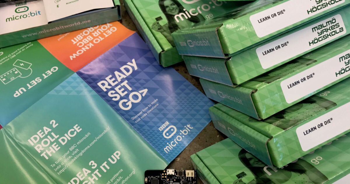 En BBC micro:bit-enhet ligger på ett bord omgiven av gröna lådor och färgglada broschyrer med programmeringsinstruktioner, inklusive fraser som READY SET GO och LEARN OR DIE.