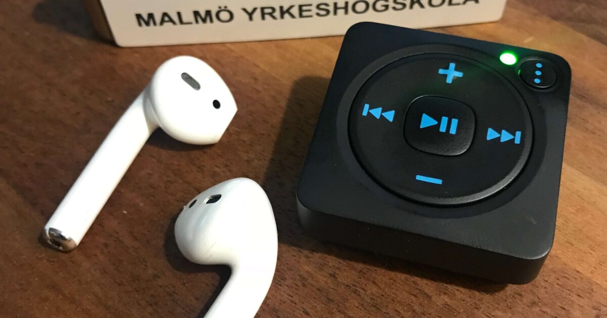 Två trådlösa hörlurar, en liten svart musikspelare med kontrollknappar och LED-lampor, och en namnbricka med texten Claes Magnusson Malmö Yrkeshögskola, står på en träyta.
