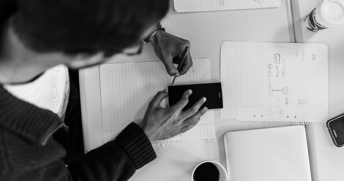 En person använder en stylus på en smartphone medan hen sitter vid ett skrivbord med en bärbar dator, en kopp kaffe och papper med flödesscheman och anteckningar. Scenen är i svartvitt.