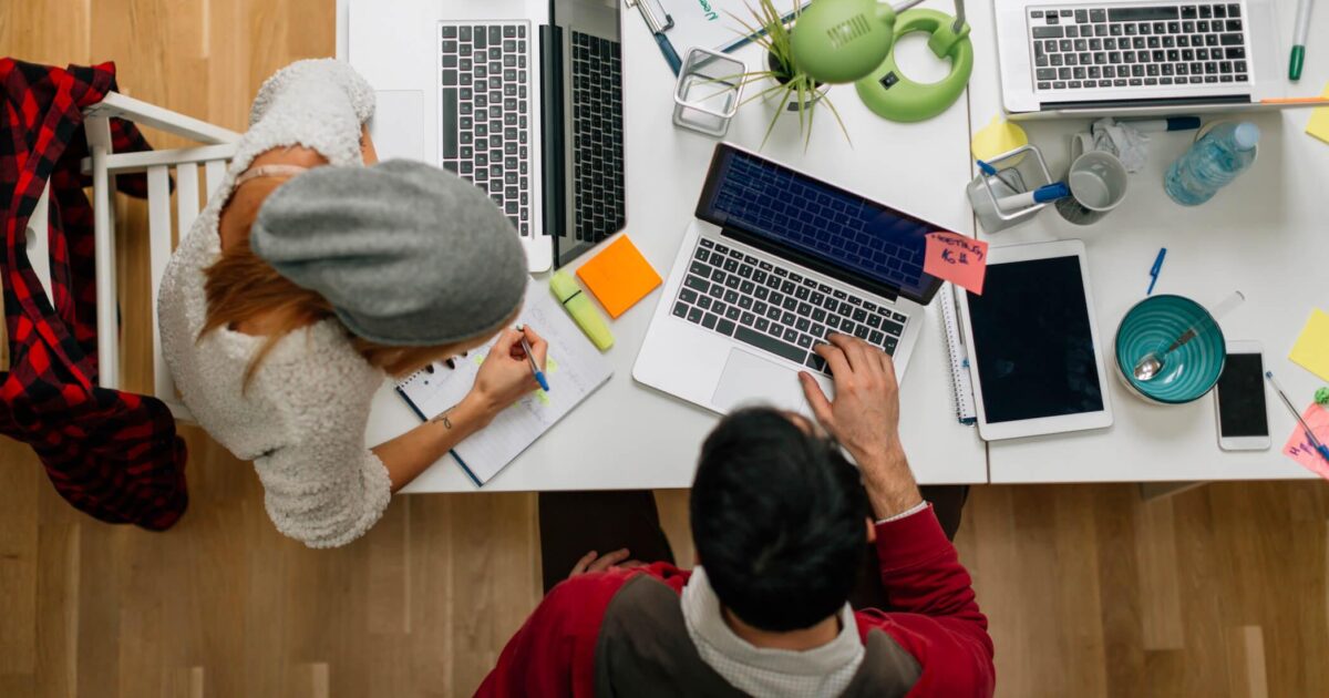 Två personer arbetar vid ett delat bord med bärbara datorer, anteckningsblock, post-it-lappar, en surfplatta, en skål och en växt. Den ena skriver i en anteckningsbok medan den andra skriver på en bärbar dator. Scenen visar en gemensam arbetsyta.