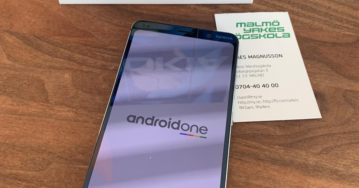 En Nokia 9 PureView-smarttelefon visar androidone-logotypen på skärmen. Bakom den finns telefonens förpackning och ett visitkort från Malmö Yrkes Högskola, allt på en träyta.
