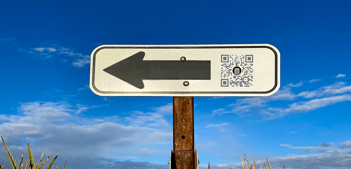 En vit skylt med en svart pil som pekar åt vänster och en QR-kod står på en rostig stolpe mot en blå himmel med moln. Grästoppar syns längst ner i bilden.