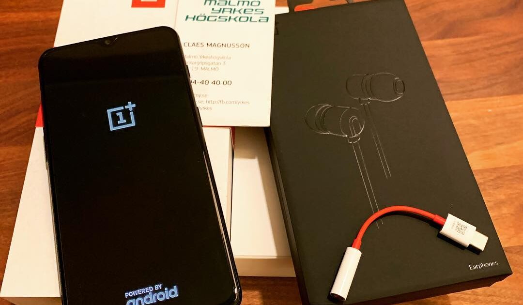 En OnePlus-smartphone som visar startskärmen ligger på sin låda bredvid hörlurar i svart förpackning, en USB-C till hörlursadapter och ett åtkomstkort från Malmö universitet på en träyta.