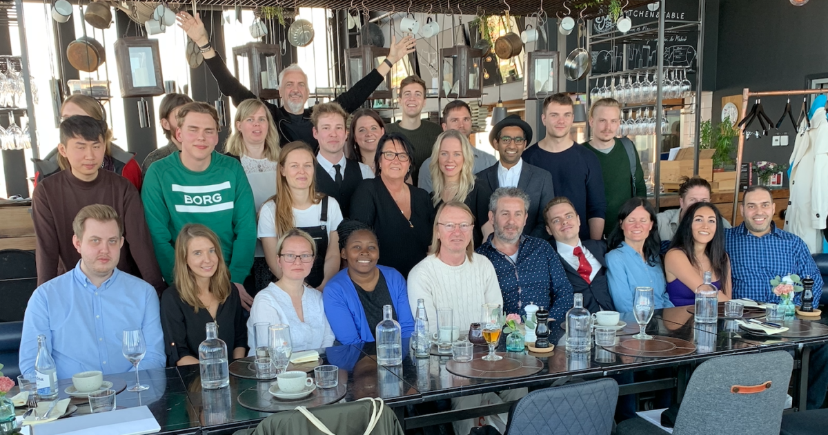 En stor grupp människor, klädda i affärsklädsel, poserar tillsammans runt ett långt matbord dukat med glas och tallrikar i en modern restaurang med hyllor och köksredskap i bakgrunden.