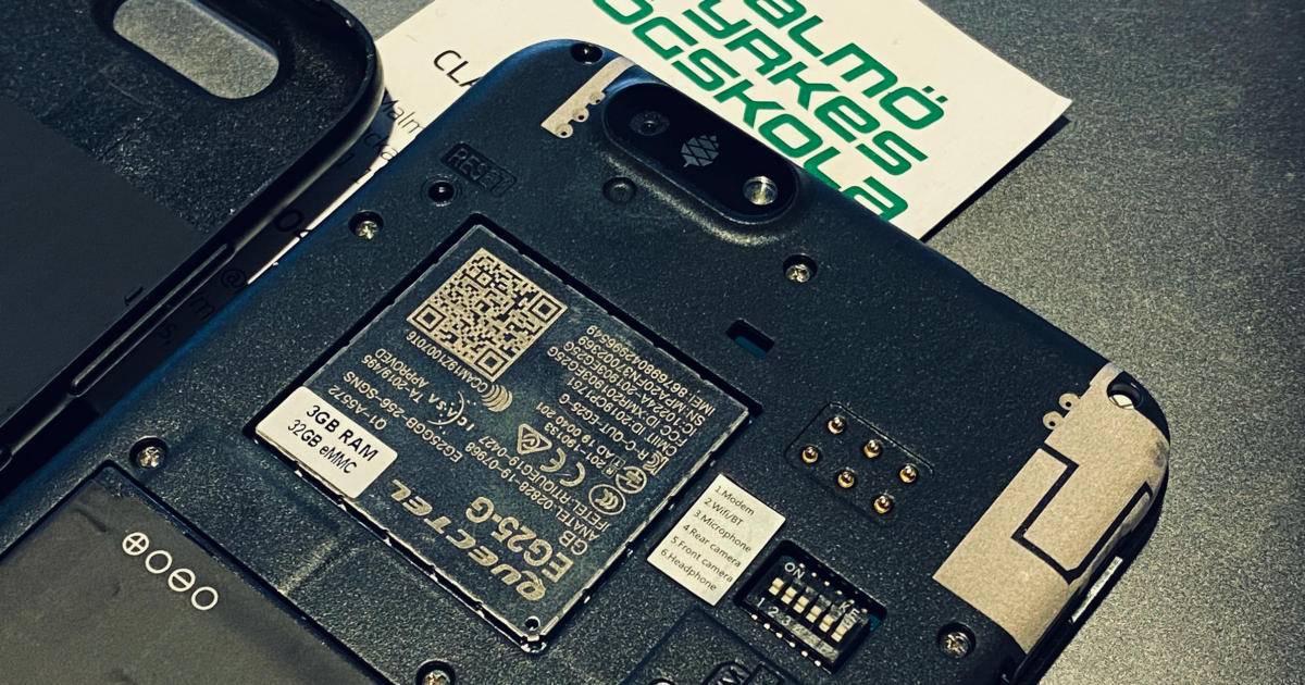 En närbild av insidan av en smartphone med bakstycket borttaget, vilket avslöjar SIM-kortplatsen, microSD-kortplatsen och batterifacket. Ett Malmö Yrkeshögskola-kort är delvis synligt under.