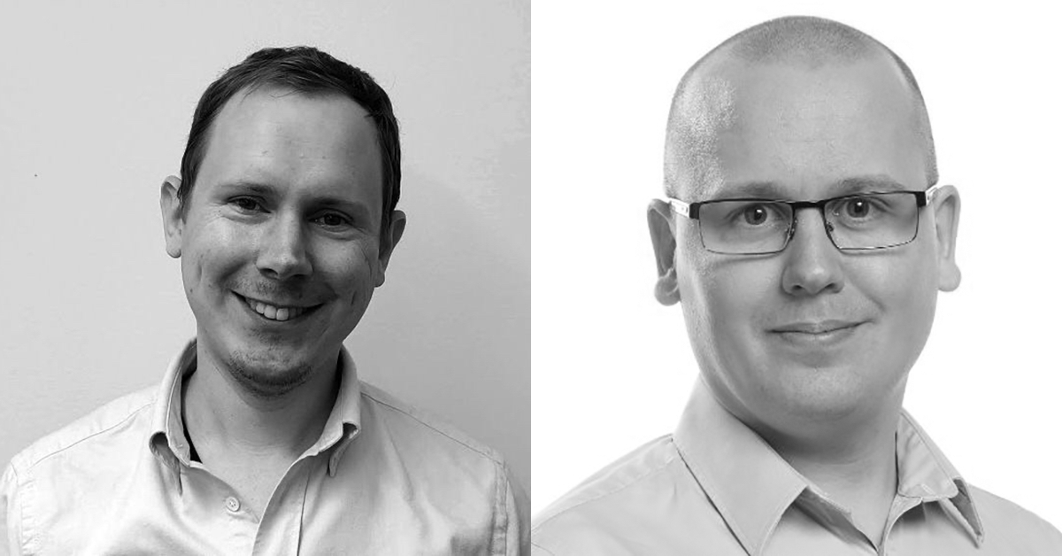 Svartvitt foto av två män sida vid sida. Mannen till vänster ler, har kort hår och bär en ljus skjorta. Mannen till höger bär glasögon, har rakat huvud och är också klädd i en ljus skjorta.