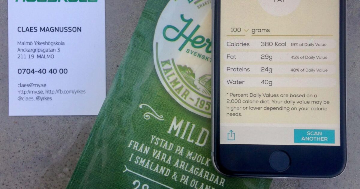 En smartphone som visar resultat från mejeriscanningar, en svart skanner, ett ostpaket märkt 150 g, 28 % fett, och ett visitkort från Malmö Yrkeshögskola på en grå yta.