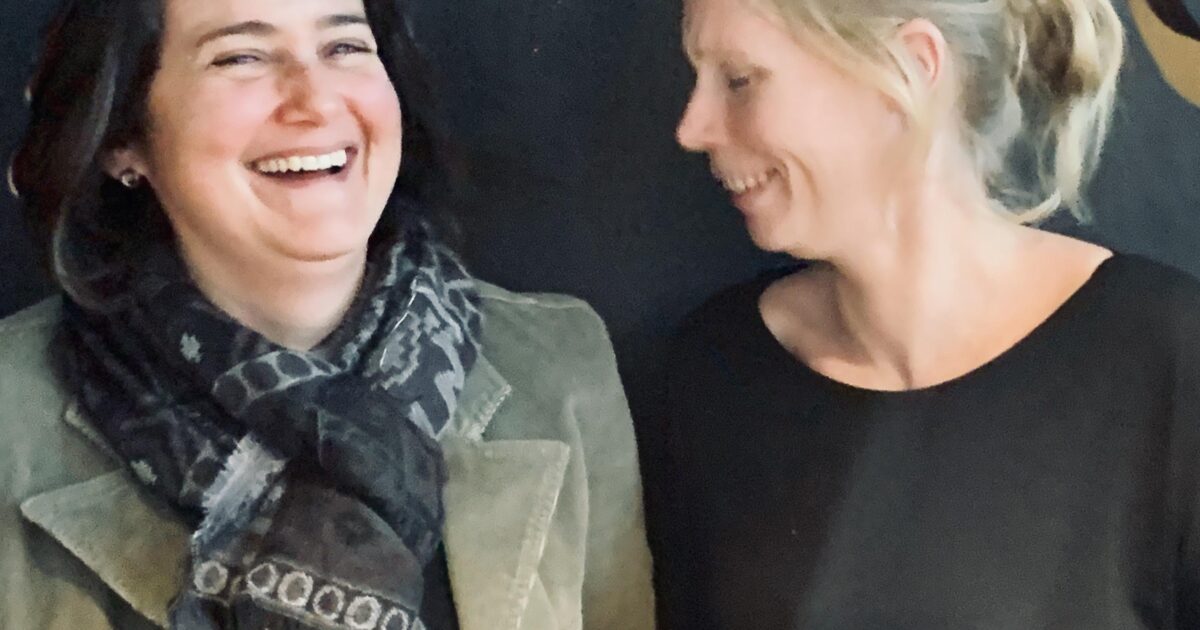 Två kvinnor står sida vid sida, ler och håller vita kuvert. Den ena bär en mönstrad halsduk och grön jacka, den andra en svart topp med blont hår uppsatt. De verkar dela ett glädjefyllt ögonblick.