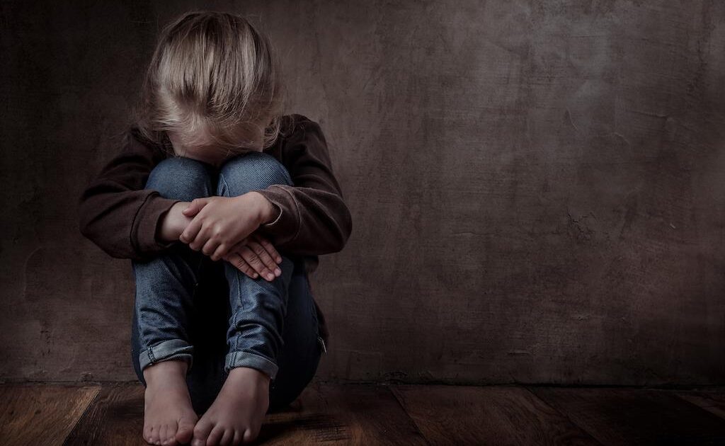 Ett litet barn sitter på ett trägolv mot en mörk vägg med huvudet nedåt och armarna runt knäna och ser ledset eller upprört ut.