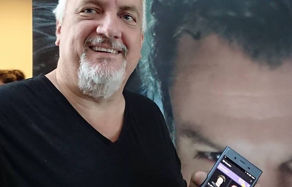 En leende man med vitt hår och skägg håller en smartphone med en app på skärmen, står framför en affisch med Sony-logotypen och en närbild av en persons ansikte.