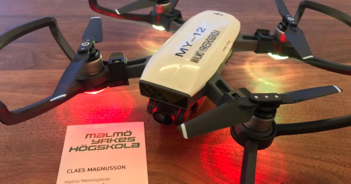En liten vit drönare med svarta propellrar står på en träyta, upplyst av röda och gröna lampor. Bredvid den ligger ett visitkort från Malmö Yrkeshögskola för Claes Magnusson.