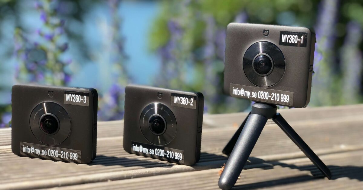 Tre svarta MY360-kameror med vidvinkelobjektiv är placerade på en träyta utomhus; två vilar platt och en är monterad på ett litet stativ, med suddig grönska och vatten i bakgrunden.