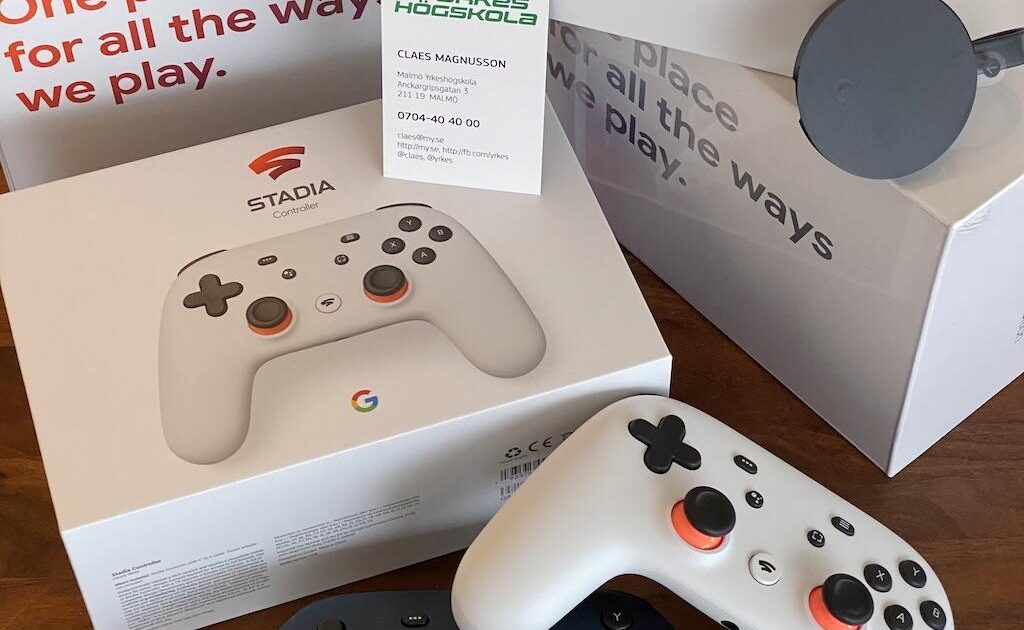 Tre Stadia-kontrollboxar, två Stadia-kontroller (en vit, en svart), en Chromecast Ultra och ett visitkort är placerade på ett träbord. Förpackningen har Stadia-logotyp.