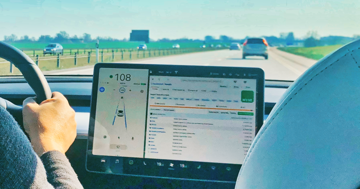 En person kör en bil på en motorväg med en digital instrumentpanel som visar hastighet, navigering och musikalternativ. Vägen framför är tydlig med några få bilar och gröna fält på båda sidor.