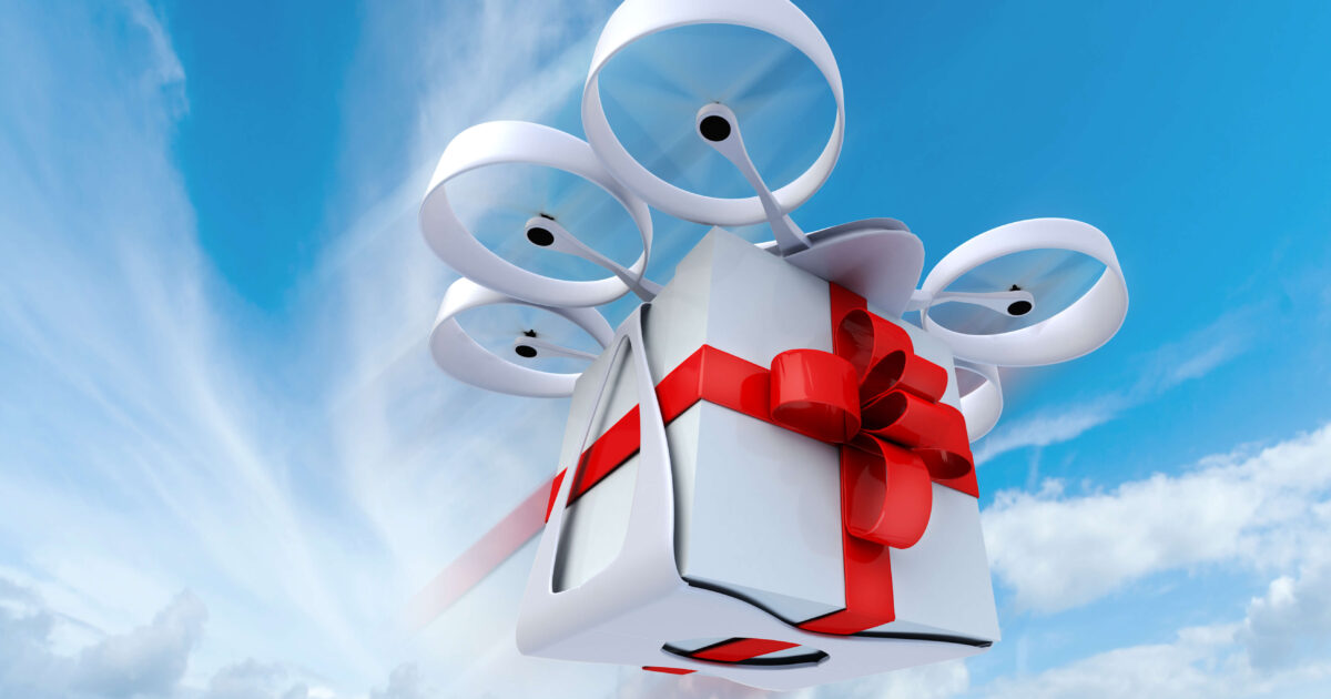 En drönare med fyra propellrar flyger på en blå himmel och bär en vit presentask inslagen med ett rött band och en rosett. Molnen syns nedanför, och lådan ser ut att vara mitt i leveransen.