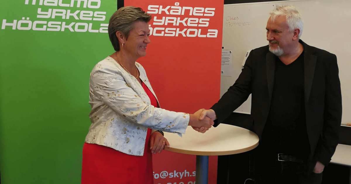 En kvinna i vitblommig kavaj och en man i svart kostym skakar hand och ler framför Malmö Yrkeshögskola och Skånes Yrkeshögskola-banderoller vid ett inomhusevenemang.