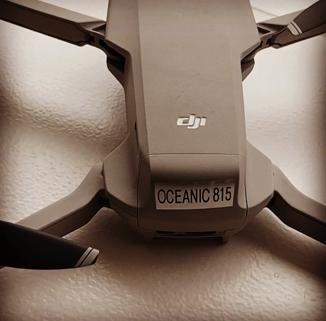 Närbild av en DJI-drönare med en etikett med texten OCEANIC 815 på kroppen, visad mot en texturerad vit bakgrund.