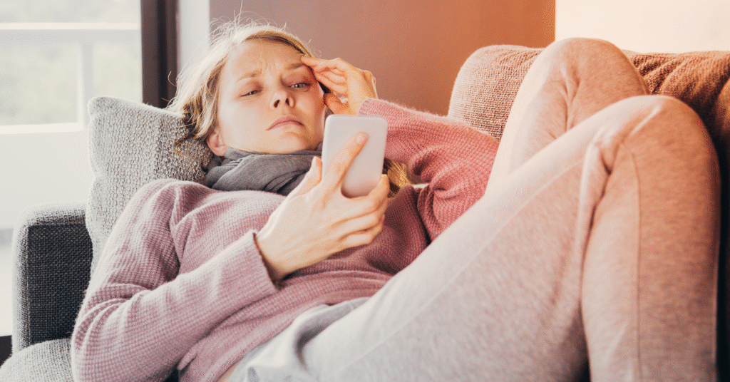 En kvinna i bekväma kläder ligger på en soffa, håller huvudet med ena handen och tittar på sin smartphone med ett bekymrat uttryck. Hon ser ut att vara orolig eller stressad medan hon läser något på skärmen.