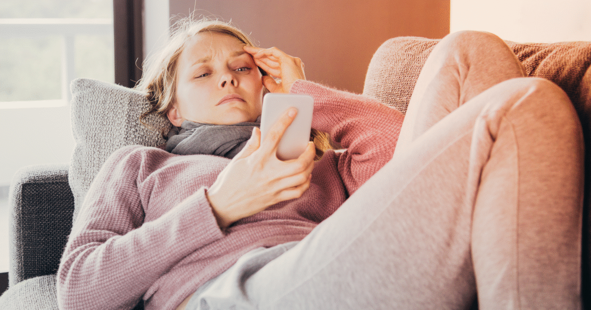 En kvinna i bekväma kläder ligger på en soffa, håller huvudet med ena handen och tittar på sin smartphone med ett bekymrat uttryck. Hon ser ut att vara orolig eller stressad medan hon läser något på skärmen.