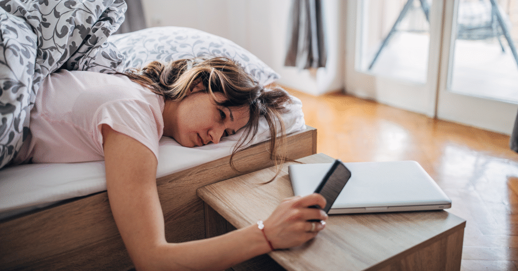 En kvinna som ligger på mage i sängen ser trött ut när hon sträcker sig efter sin telefon på ett nattduksbord, bredvid en stängd laptop. Morgonljus kommer in genom närliggande glasdörrar.