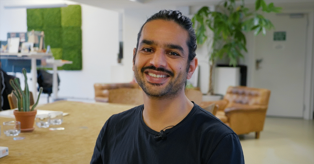 En man med långt mörkt hår som är bakåtkammat och skägg ler mot kameran i ett ljust, modernt kontor med växter, solbruna stolar och ett stort bord i bakgrunden.