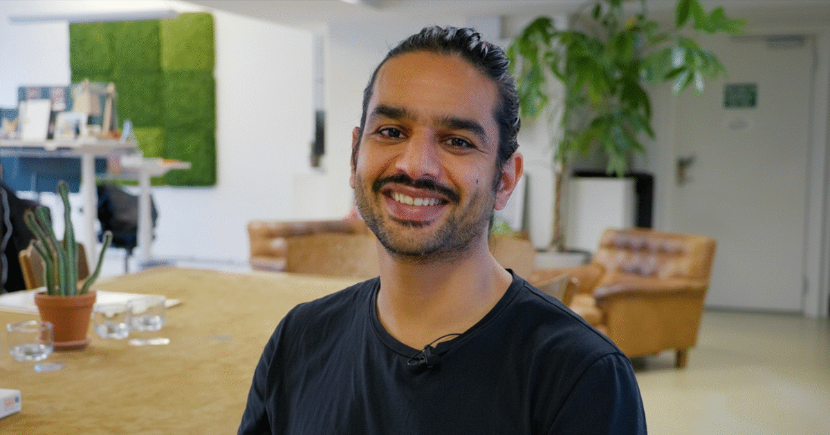 En man med långt mörkt hår som är bakåtkammat och skägg ler mot kameran i ett ljust, modernt kontor med växter, solbruna stolar och ett stort bord i bakgrunden.