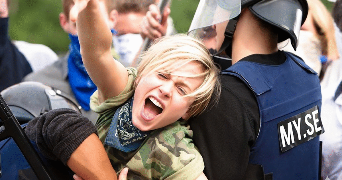 En person med kort blont hår och en bandana runt halsen skriker medan han hålls fast av en säkerhetsvakt i kravallutrustning under en demonstration.