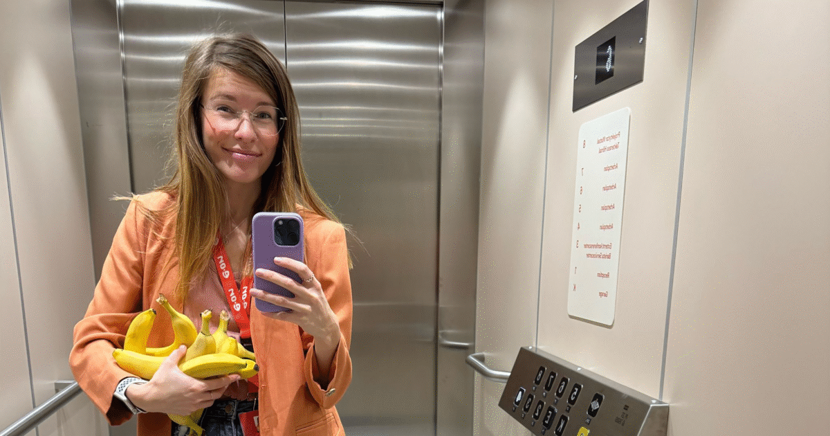 En kvinna i orange kavaj står i en hiss med flera bananer i handen och tar en selfie med sin telefon. Hon bär glasögon och ett rött nyckelband och hissens väggar är av metall med synliga knappar och en våningspanel.