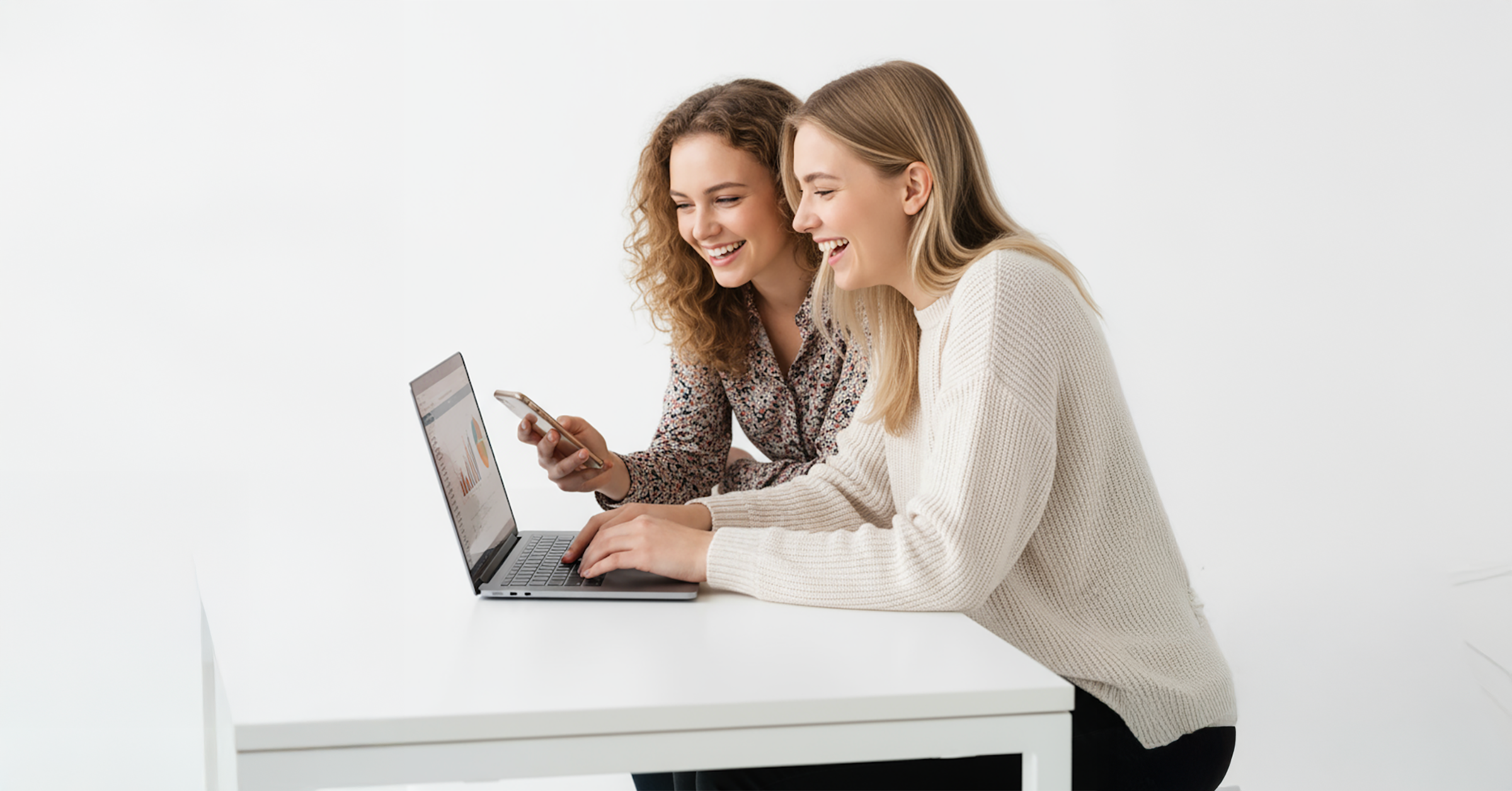 Två unga kvinnor sitter vid ett vitt bord, ler och tittar på en laptopskärm. Den ena skriver på den bärbara datorn medan den andra håller i en smartphone, båda verkar engagerade och glada i en ljus, minimalistisk miljö.