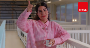 En kvinna i rosa randig skjorta ler och gör tummen upp medan hon håller i en mugg, stående i en ljus inomhusmiljö. MY.SE-logotypen syns i det övre högra hörnet.