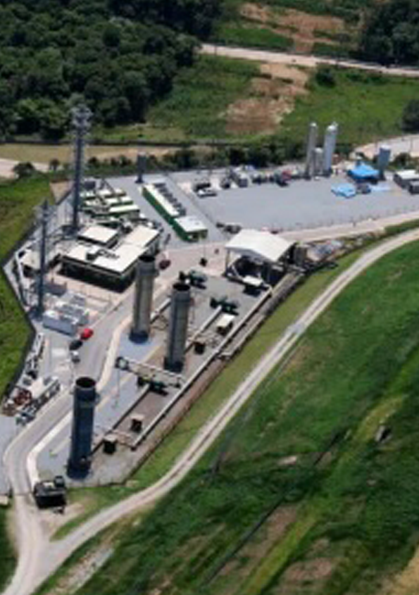 Flygfoto över en industrianläggning med flera höga cylindriska strukturer, byggnader och fordon, omgiven av grönska och en slingrande väg.