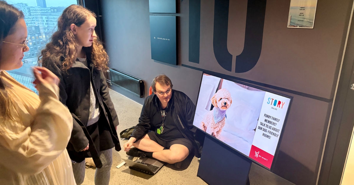 Tre personer tittar på en digital skärm som visar en bild av en hund och text. En person sitter på knä bredvid skärmen, medan de andra två står i närheten. Miljön ser ut att vara inomhus med stadsbyggnader som syns genom ett fönster.