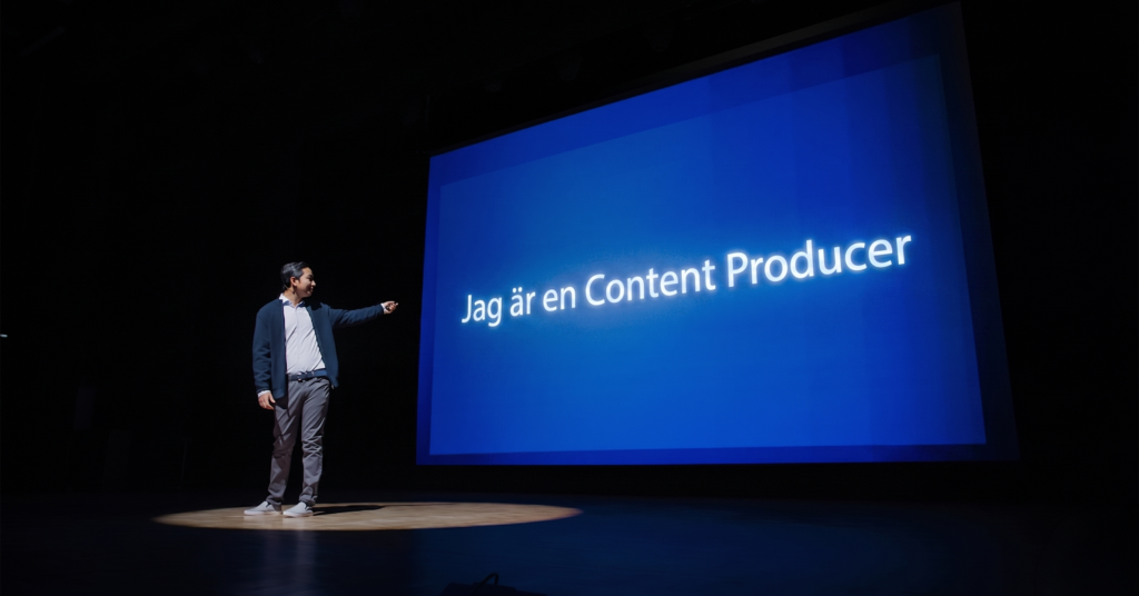 En man står på en scen i strålkastarljus och pekar på en stor blå skärm där det står Jag är en Content Producer, vilket betyder Jag är en innehållsproducent.