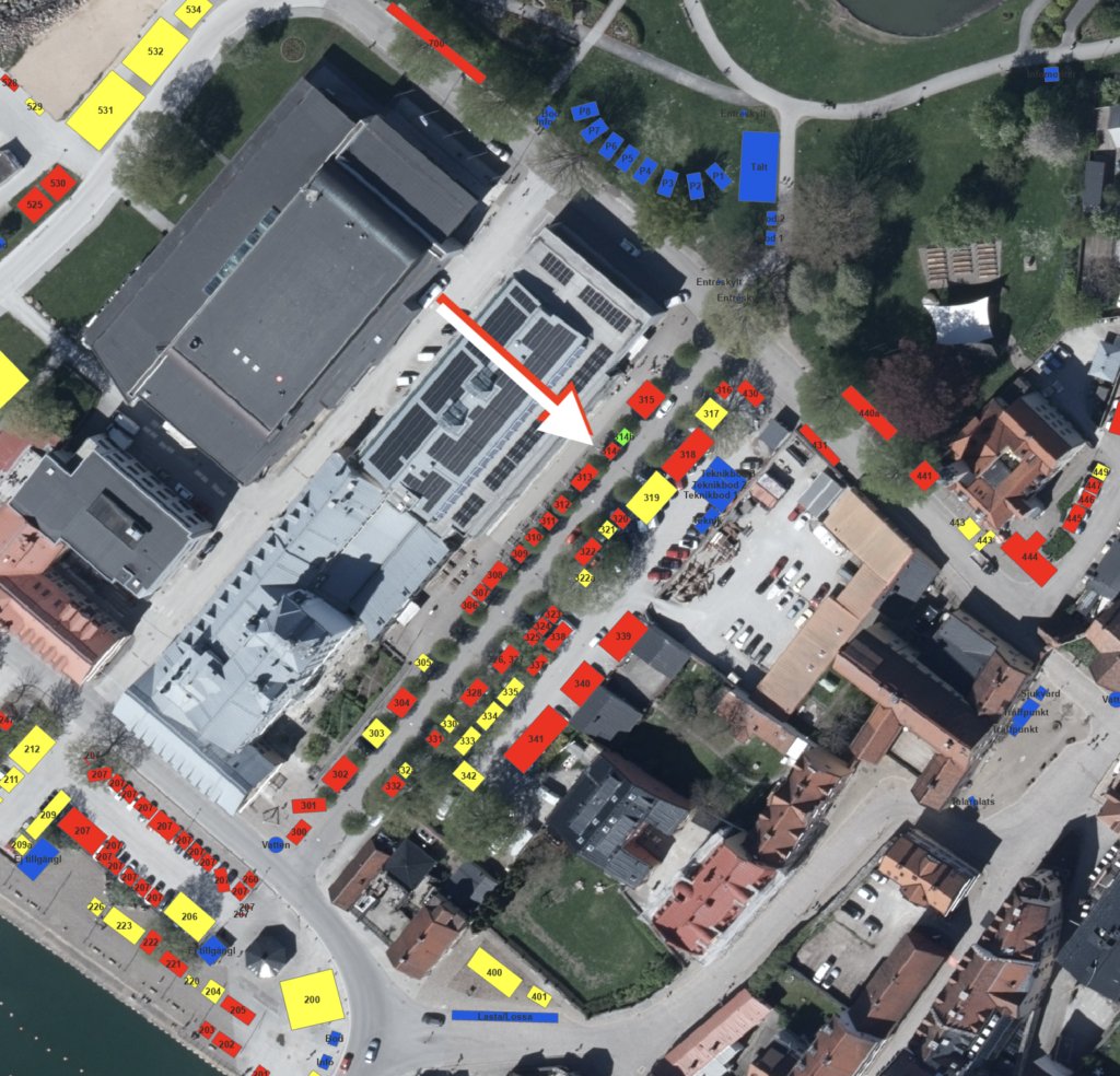 Flygbild över ett kvarter med byggnader, parkeringsplatser och vägar; olika parkeringsplatser är markerade med röda, blå och gula rutor med siffror, och en stor vit pil visar en väg genom den centrala parkeringsplatsen.