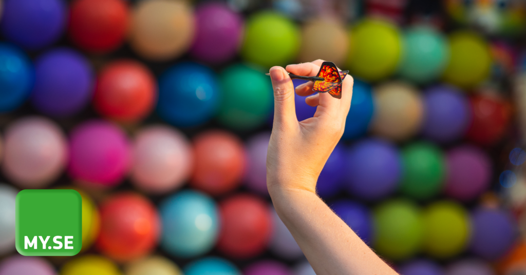 En hand håller en pil framför en färgglad vägg av ballonger vid en spelbås på en nöjespark. I det nedre vänstra hörnet syns en grön fyrkant med texten MY.SE.