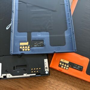 theotherhalf2 Tre Kindle Jolla-baksidor – en blå, en orange och en svart – med synliga anslutningar och kretskort på ett träbord.