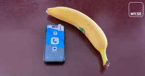 En smartphone med ikoner för samtal och chatt ligger bredvid en mogen banan på ett vinrött bord. I det övre högra hörnet syns en vit MY.SE-logotyp i en rundad fyrkant.